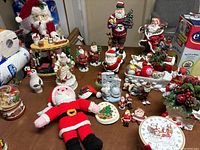 Tabletop display of assorted Christmas decorations including several Santa Claus figurines, a hand-knit Santa doll, small holiday plates, and various small ornaments and decorative pieces.