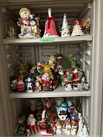 Photo showing three shelves filled with various Christmas-themed figurines, including Santa Claus, snowmen, angels, reindeer, candle holders, and decorative ornaments.