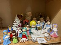 Photo shows ceramic nativity figurines including Mary, Joseph, baby Jesus, wise men, angels, sheep, a white ceramic Christmas tree, and two small wrapped silver decorative items.