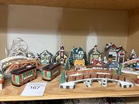 Front view of ceramic Christmas village houses, bridges, pine trees, and train cars arranged on wooden shelf.