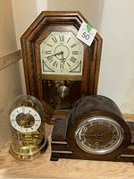 Wooden octagonal wall clock with pendulum, Kundo anniversary mantel clock with glass dome, and wooden mantel clock with round dial displayed together on wooden surface