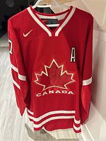 Front view showing red jersey with 'Canada' maple leaf crest, Nike swoosh, alternate captain 'A' patch