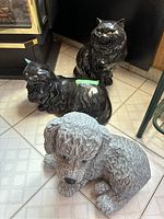 Two black ceramic cat figurines and one gray ceramic dog figurine arranged on tiled floor with light pattern. Cats glossy and dog textured.
