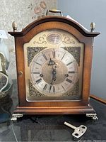 Front view of the wooden Tempus Fugit mantel clock with brass decorative elements and key in front.