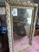 Full view of rectangular mirror with ornate gold-tone frame resting against furniture on a carpeted floor.