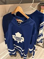 Two blue Toronto Maple Leafs hockey jerseys hanging on hangers showing front and side views with logos and striping details.