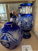 Two vintage cobalt blue glass vases displayed together in a cabinet. One round and one taller with flared rim, both decorated with silver cut glass patterns.