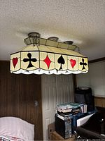 Ceiling-mounted stained glass light fixture with poker card suit pattern along the sides, hanging in a basement room with wood paneling and door.