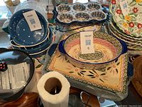 Shows set of colorful ceramic bakeware including oval casserole dishes, rectangular casserole dish, blue polka dotted dish, two serving trays, and a wire rack.