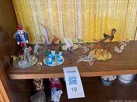 Shelf display showing multiple blown glass animal and fantasy figurines in various colors including blue, clear, amber, and red, arranged on wooden shelf.