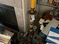 Tall candle holder made of brass and marble with a yellow candle on top, positioned near a fireplace and cluttered room items