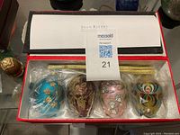 Photo of a red box containing four individually wrapped glass Christmas ornaments by Joan Rivers, displayed on a table with other decorative items.