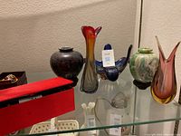 Blown glass vases and decorative items displayed on glass shelves, various colors and shapes including black, green/white marbled, amber/red, and blue glass bird sculpture.