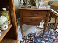 Front view of one wooden end table showing two drawers with ornate brass handles, placed on a colorful patterned rug.