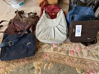 12 purses displayed on a floral carpet. Various colors and styles, mostly leather and some fabric bags.