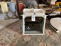 White countertop rotisserie cooker with glass door front and removable drip trays on top, placed on a carpeted floor with other household items nearby.