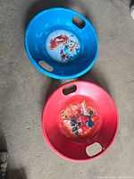 Photo showing two round plastic kids snow sledding boards, one blue with a snowman and snowboarder cartoon, one red with cartoon characters, both have hand grips.
