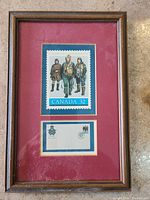 Framed stamp and emblem with red matting and wooden frame on floor.