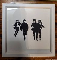 Framed wall art showing black silhouettes of The Beatles walking, placed on wooden surface with visible reflections on glass.