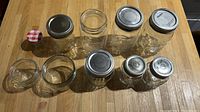 10 assorted glass jars placed on a wooden surface. 7 jars have metal lids, 3 jars without lids. Sizes range from small to medium height.