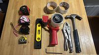 Top-down image displaying all tools: four measuring tapes arranged on left, yellow spirit level, red tape dispenser with tape, metal Allen key, silver vice grip, and claw hammer with black handle on wooden table