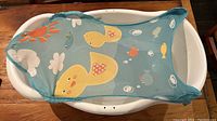 Top view of white plastic baby bathtub containing blue mesh sling with duck and fish patterns designed for infant support.