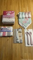 Full lot displayed on wooden floor: 6 pack cotton receiving blankets, 6 bibs with attached socks in pastel shades, 6 pastel washcloths, pack of 2 pink baby spoons, and packaged linen.