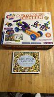 Photo showing box of STEM Automobile Engineer Kit for ages 3-6 with colorful toy vehicles on cover, and the children's book "Each Peach Pear Plum" with illustrated cover by Janet and Allan Ahlberg.