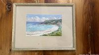 Full view of the framed painting showing the scenic beach landscape with blue ocean, sky, greenery, and sandy shore.
