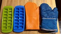 Photo shows two silicone ice trays (green and blue) and two oven mitts (orange silicone and blue fabric) on a wooden floor.