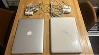 Top view of two MacBook laptops side by side with respective charger cables on a wood table: the older white 2004 MacBook on the right and the newer silver 2017 MacBook on the left.