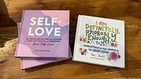 Three books, two "Self Love" in pink cover and one "I am Definitely, Probably Enough" with white and colorful text cover laid out on wooden surface.