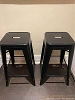 Two black metal square seat counter stools placed side by side against beige wall on dark floor, showing overall condition and shape.
