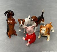 Photo shows five mini Beswick ceramic figurines: four dogs including a brown dachshund standing on hind legs, a white and brown Jack Russell terrier, a tricolor beagle, a tan and white bulldog, and a pink pig in red sweater sitting. All placed on gray fabric background.
