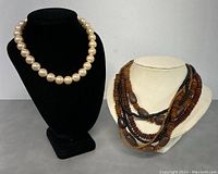Front view of vintage pearl necklace on black display and multiple strand wooden bead necklaces on cream display.