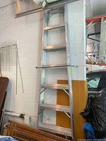 Photo of an aluminum ladder stored vertically against a white cinderblock wall in a garage space, showing the full ladder with several rungs visible.