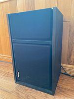 Front angle view of a black Bose speaker placed on wooden floor against wood paneling.