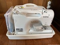 Front right view of white Brother sewing machine in base of plastic carrying case showing machine body, free arm and manuals