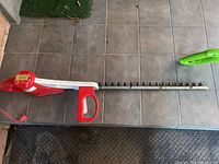 Electric hedge trimmer lying on tiled floor showing full length with the red handle, cord, and blade