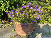 Front view of fibreglass planter with purple flowering plant. Shows decorative floral patterns and missing handle.