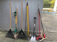 Photo showing 9 gardening and cleaning tools standing against gray wall, including rakes, shovels, hoes, and brooms.