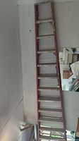 Image showing a 12ft red folding aluminum ladder leaning against a wall inside a garage or indoor space with visible wear and paint marks.