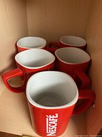 Five red ceramic mugs with large white Nescafé logo, placed inside a wooden cabinet, front view showing inside of one mug and logo on the others.