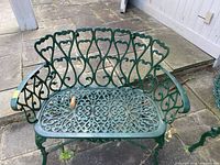 Green cast aluminum bench with decorative openwork on seat and backrest, showing some leaves and debris on the seat. Outdoor setting on stone patio.