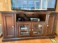 Front view of the dark wood TV console with a Panasonic TV on top, showing two solid doors on the sides and glass front double doors in the center with internal shelves and items inside.