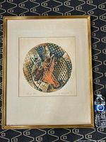 Framed abstract colored etching by Jaroslav Hovadik placed on dark patterned surface with water bottle for scale.