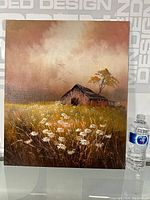 Full view of the oil painting leaning upright against a backdrop with branding. Shows the barn, tree, and flowers. A water bottle is placed next to it for scale.