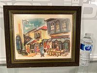 Full view of framed watercolour painting with bottle for scale, showing a street market scene with people, market stalls, and traditional buildings.