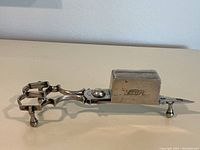 Side view of antique silver candle snuffer showing hexagonal handle, three feet, and hinged box with engraved marks.