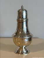 Full front view of a polished sterling silver sugar shaker showing its tapered, bulbous body with decorative ridges topped by a pierced shaker lid with a finial.